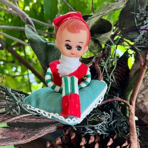 Puede incluir: Una pequeña figura de elfo con cara de plástico moldeado, rasgos pintados y un gorro rojo. El elfo viste una parte superior roja y blanca con mangas a rayas verdes y blancas, y está sentado sobre un cojín de fieltro turquesa con encaje.