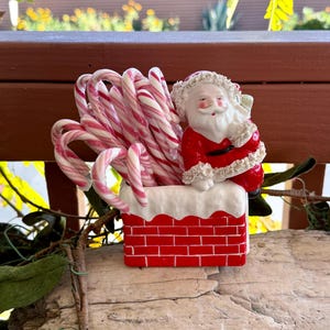 May include: A Christmas ornament depicting a red brick chimney filled with pink and white striped candy canes. A Santa Claus figure in a red suit and white beard sits on top, creating a festive holiday display.
