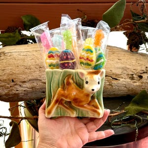 Puede incluir: Una maceta de cerámica con un diseño de cervatillo, llena de caramelos en forma de huevo de Pascua envueltos individualmente. La maceta es de color verde claro con un cervatillo marrón. Los caramelos son coloridos, con tonos amarillos, azules, rosas y verdes.