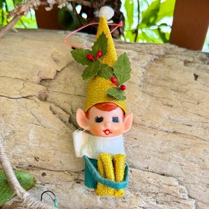 Puede incluir: Adorno navideño vintage de elfo con gorro amarillo tejido, hojas verdes y bayas rojas. El elfo tiene una cara pintada, cuello blanco y atuendo verde y amarillo. El adorno se exhibe sobre un trozo de madera.