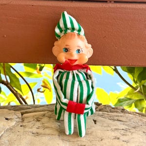 Puede incluir: Una pequeña muñeca elfo con cara de plástico, ojos azules pintados y boca roja. La muñeca viste un conjunto de tela a rayas verdes y blancas con un gorro puntiagudo a juego, un cuello rojo y puños rojos en las mangas de la prenda.