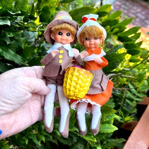May include: Two small dolls dressed in pilgrim-style felt clothing. One doll wears a brown hat and jacket with yellow buttons, while the other wears an orange dress with a white bonnet. They hold a small yellow plastic basket between them.