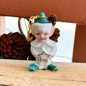 May include: A vintage ceramic Christmas ornament in the shape of a smiling elf. The elf wears a white outfit with a green hat and shoes. A gold star is on the front. The ornament is suspended by a gold string.