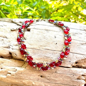 May include: A necklace featuring a series of red, leaf-shaped glass stones alternating with small, iridescent, round crystals set in a gold-tone metal frame. The design includes a metal chain with a small charm at the end of the clasp mechanism.