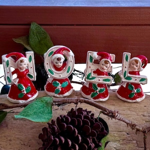 May include: Ceramic Christmas figurines spelling "HOLE" in red, white, and green. Each letter features a figurine wearing a Santa hat and red dress. The figurines are on a wooden surface with greenery and a pinecone.