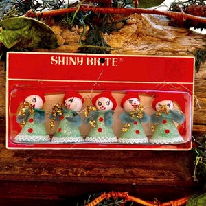 Puede incluir: Un conjunto de cinco adornos navideños Shiny Brite en su empaque rojo original. Cada adorno presenta una pequeña figura con gorro rojo, cara blanca y un vestido verde claro brillante con botones rojos, encaje blanco y detalles dorados.