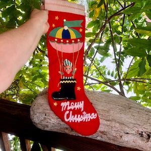 May include: A red felt Christmas stocking featuring a decorative hot air balloon applique with a small elf figure inside a black boot basket. The stocking includes gold string accents, yellow star appliques, and the white embroidered text Merry Christmas.