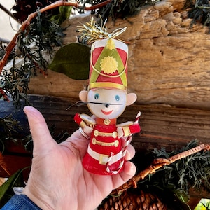 May include: A vintage Christmas ornament in the shape of a toy soldier mouse. The ornament is red, white, and gold, with a drum and a candy cane. The mouse has a white face, blue eyes, and black whiskers. The soldier's hat is red and green.
