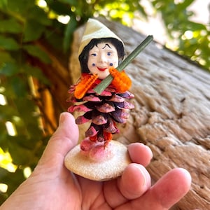 May include: A small figurine crafted from a pinecone with a painted face, wearing a cream-colored pointed hat. The figure has orange pipe cleaner arms, holds a thin green stick, and stands on a circular, light-colored base with pink accents.