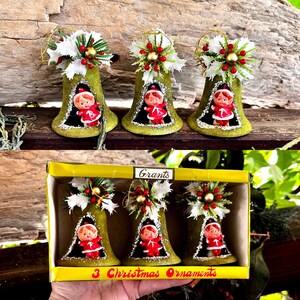 May include: Three bell-shaped Christmas ornaments with a green flocked texture, each featuring a small elf figure in a red suit inside. The top of each bell is decorated with white holly leaves, red berries, and a gold bead. The box is labelled Grants.