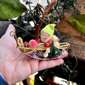 May include: A vintage Christmas ornament featuring a green-hatted gnome riding in a gold-wire sleigh with a pink ball and green pine branches.