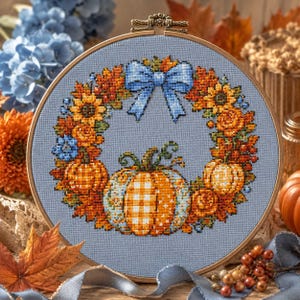 Puede incluir: Un bastidor de bordado circular de madera que contiene un diseño de punto de cruz sobre tela azul claro. El diseño presenta una corona de calabazas naranjas, girasoles y hojas de otoño, centrada con una calabaza grande y un lazo azul claro.