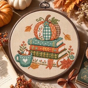 May include: A circular wooden embroidery hoop containing a cross-stitch design of a pumpkin resting on a stack of three books, next to a teal mug with a heart, surrounded by autumn leaves and berries on a light beige fabric background.