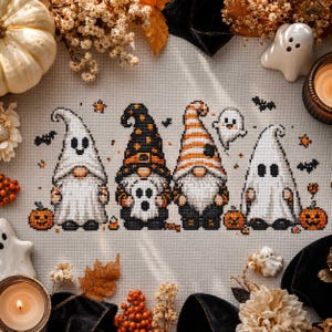 以下が含まれることがあります： 明るい布地に施されたクロスステッチの図案で、ハロウィンの仮装をした4体のノームが描かれています。2体はゴーストの衣装を着ており、1体は星柄の黒い帽子、もう1体はオレンジ色の縞模様の帽子をかぶっています。周囲には小さなカボチャ、コウモリ、ゴーストが配置されています。