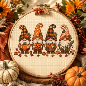 May include: A circular wooden embroidery hoop containing a cross-stitch pattern of four gnomes wearing patterned hats in shades of orange, brown, and black. Each gnome holds a pumpkin, with scattered autumn leaves and small pumpkins at their feet.