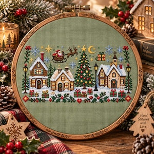 May include: A circular wooden embroidery hoop containing a cross-stitch pattern on sage green fabric. The design features a snowy village scene with houses, a decorated Christmas tree, street lamps, wrapped gifts, and Santa in a sleigh.