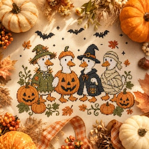 Può includere: Un ricamo a punto croce su tessuto beige raffigurante quattro anatre in costume: una con cappello verde, una in costume da zucca, una con cappello da strega nero e una da mummia. Il disegno include pipistrelli neri, zucche e foglie autunnali.