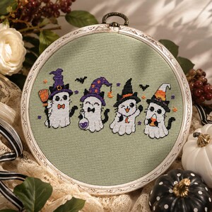 May include: A circular embroidery hoop containing a cross-stitch pattern on sage green fabric. The design features four white ghost-like figures wearing colorful witch hats, holding items including a broom, a potion bottle, and candy corn.