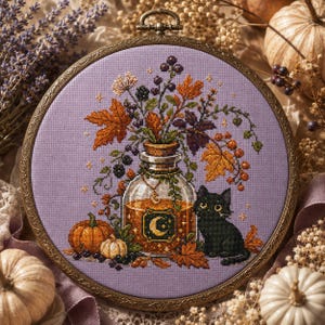 May include: A circular embroidery hoop containing a cross-stitch design on lavender fabric. The pattern features a glass bottle filled with amber liquid, a black cat, orange autumn leaves, berries, and a pumpkin, all framed in antique bronze.