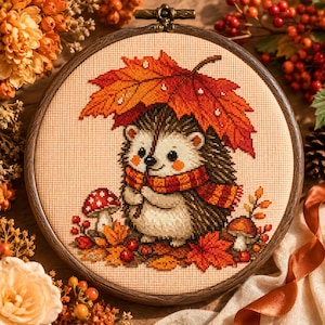 May include: A circular embroidery hoop containing a cross-stitch pattern of a hedgehog wearing a red and orange striped scarf, holding a large orange maple leaf over its head, surrounded by red mushrooms and autumn leaves on beige fabric.