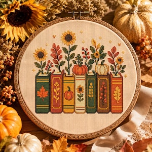 May include: A circular embroidery hoop containing a cross-stitch pattern of seven colorful books arranged in a row. Each book features a unique botanical design, including sunflowers, pumpkins, and autumn leaves, stitched in shades of green, red, orange, and yellow on a light beige fabric background.