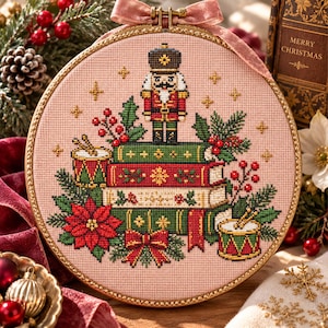 May include: A circular embroidery hoop featuring a cross-stitch design on light pink fabric. The pattern depicts a nutcracker standing on a stack of three books, surrounded by drums, poinsettias, holly, and small gold stars.