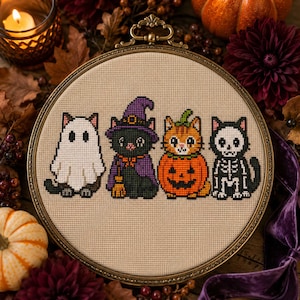 May include: A circular embroidery hoop containing a cross-stitch pattern on beige fabric. The design features four cats in costumes: one as a ghost, one as a witch with a purple hat, one inside a pumpkin, and one wearing a white skeleton suit.