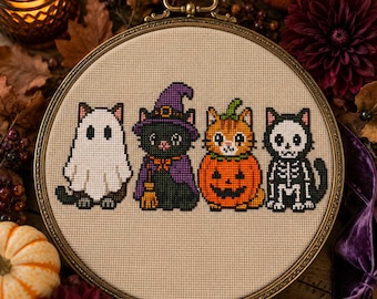 Puede incluir: Un bastidor de bordado circular con un patrón de punto de cruz sobre tela beige. El diseño muestra cuatro gatos disfrazados: uno de fantasma, uno de bruja con sombrero morado, uno dentro de una calabaza y otro con un traje de esqueleto blanco.