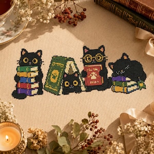 May include: A cross-stitch embroidery on beige fabric featuring four black cats. One cat holds a stack of colorful books, another peeks from behind a green book, a third wears round glasses while holding a red book, and the fourth sleeps on books.