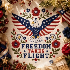May include: A cross-stitch embroidery on white fabric featuring a bald eagle with wings spread, colored in red, white, and blue. Below the eagle, the text reads FREEDOM TAKES FLIGHT in blue and red block letters, surrounded by floral motifs.