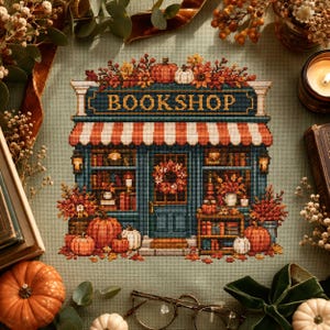 May include: A cross-stitch embroidery on sage green fabric depicting a bookshop storefront with a striped awning, the word BOOKSHOP in yellow letters, a wreath on the door, shelves filled with books, and pumpkins in autumn colors.