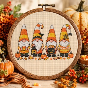 Puede incluir: Un bastidor de bordado circular de madera que contiene un patrón de punto de cruz de cuatro gnomos. Los gnomos llevan gorros en colores naranja, amarillo y blanco. Están representados con calabazas, un farol y un caldero negro.