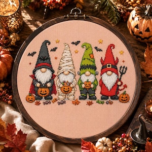 May include: A circular wooden embroidery hoop containing a cross-stitch pattern of four Halloween gnomes on a light peach fabric. The gnomes are dressed as a vampire, a mummy, Frankenstein's monster, and a devil, each holding a small pumpkin.