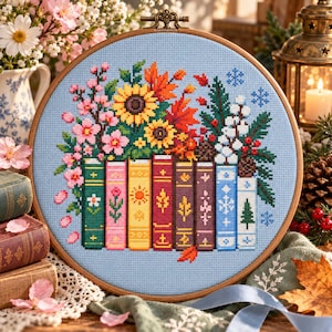 May include: A circular wooden embroidery hoop containing a cross-stitch design on light blue fabric. The pattern features a row of colorful books topped with seasonal floral arrangements, including sunflowers, pink blossoms, pine branches, and berries.