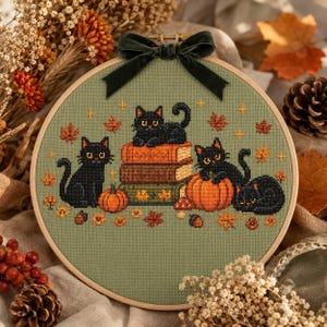 May include: A circular wooden embroidery hoop containing a cross-stitch pattern on sage green fabric. The design features four black cats, a stack of three books, pumpkins, mushrooms, and scattered autumn leaves, topped with a dark green velvet bow.
