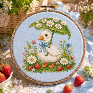 May include: A cross-stitch embroidery in a wooden hoop featuring a white duckling with an orange beak and a blue bow tie, holding a large green leaf umbrella. The duckling is surrounded by white daisies, red strawberries, and blue flowers.