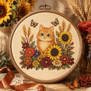 May include: A circular wooden embroidery hoop containing a cross-stitch design of an orange tabby cat surrounded by sunflowers, wheat stalks, and autumn-coloured flowers. Two small butterflies are stitched above the cat on cream-coloured fabric.