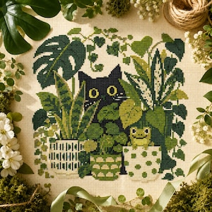 Puede incluir: Un bordado de punto de cruz sobre tela beige que muestra un gato negro asomándose entre varias plantas verdes, incluyendo una monstera y una sansevieria, junto a una pequeña rana verde sentada en una maceta de lunares blancos entre follaje.