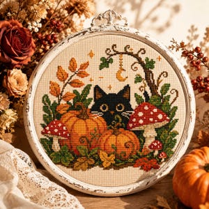 Könnte beinhalten: Eine kreisförmige Kreuzstichstickerei in einem weißen Rahmen mit einer schwarzen Katze, umgeben von orangefarbenen Kürbissen, rot-weiß gepunkteten Pilzen, grünem Blattwerk und Herbstblättern sowie einem goldenen Halbmond am Ast.