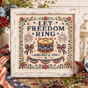 May include: A square cross-stitch sampler in a distressed white wooden frame featuring the text LET FREEDOM RING and AMERICA 250. The design includes a central drum with drumsticks, fireworks, bows, and floral borders in red, white, and blue.