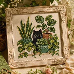 Pode incluir: Um bordado em ponto cruz numa moldura decorativa de cor creme envelhecida. O design apresenta um gato preto com grandes olhos amarelos sentado entre várias plantas de interior em vasos, incluindo uma sansevieria e folhagem pendente.
