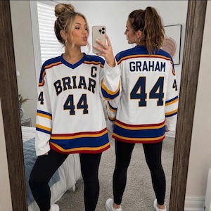 Könnte beinhalten: Ein weißes Eishockeytrikot mit blauen und gelben Streifen an den Ärmeln und am Saum. Die Vorderseite zeigt den Schriftzug BRIAR, die Nummer 44 und ein C. Die Rückseite zeigt den Namen GRAHAM und die Nummer 44 in dunkelblauer Schrift mit gelben Rändern.