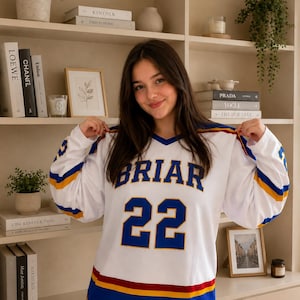 May include: A white long-sleeve jersey featuring the word BRIAR in blue letters with yellow outlines above the number 22 in large blue font. The sleeves and hem have blue and yellow stripes, with a red accent stripe near the bottom hem.
