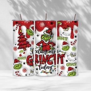 May include: A white stainless steel tumbler with a red drip pattern at the top. It features a green character in a Santa hat, a red Christmas tree, candy canes, snowflakes, and the text Feeling extra GRINCHY today in red and black.