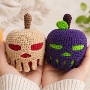May include: Two crocheted apple-shaped figures with skull-like faces. One is beige with red eyes and mouth, the other is purple with bright green eyes and mouth. Both have brown stems, and the purple one features a small green leaf on its stem.