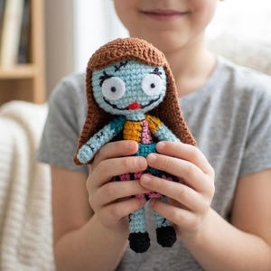 Könnte beinhalten: Eine handgefertigte Häkelpuppe mit hellblauem Gesicht, großen weißen runden Augen, einem aufgestickten schwarzen Mund und einer kleinen roten Nase. Die Puppe hat langes braunes Garnhaar und trägt ein Kleid im Patchwork-Stil mit blauen, gelben und rosa Abschnitten.