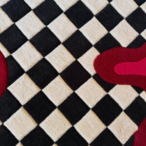 May include: A tufted rug featuring a repeating black and off-white checkered pattern. Overlaid on the right side is a large, organic, fluid shape in two shades of red, with a smaller portion of a similar red shape visible on the far left edge.