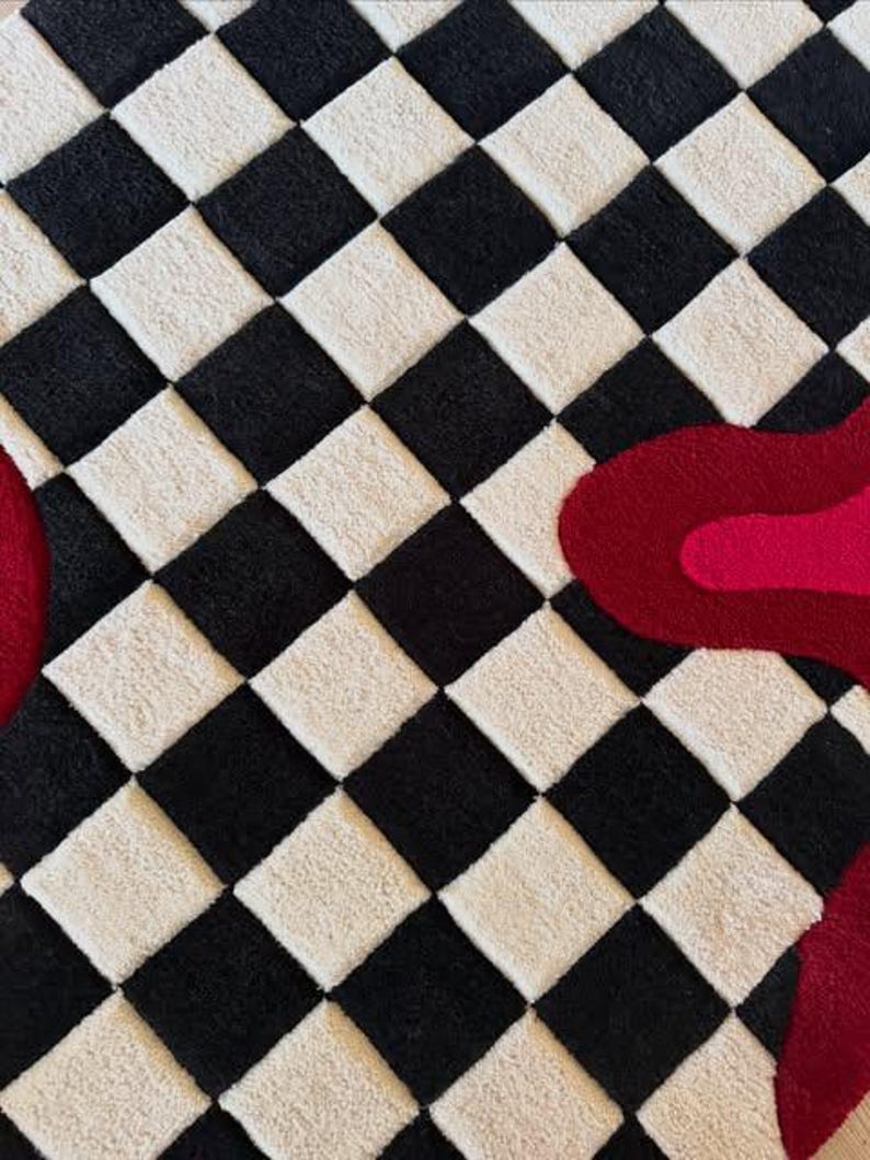 May include: A tufted rug featuring a repeating black and off-white checkered pattern. Overlaid on the right side is a large, organic, fluid shape in two shades of red, with a smaller portion of a similar red shape visible on the far left edge.
