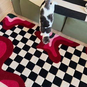 Hand tufted checkerboard rug featuring bold black and white checkered pattern with abstract pink, red, blush and burgundy wavy shapes. Modern handmade wool area rug designed for contemporary living rooms, bedrooms, apartments, dorm rooms and creative interiors. Unique dopamine decor rug with artistic retro aesthetic, colorful statement design and plush texture. Perfect for modern home decor, eclectic interiors, maximalist styling, trendy room makeover, luxury carpet styling, aesthetic room decor