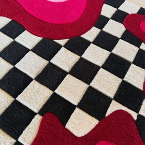 May include: A tufted rug featuring a black and white checkered pattern contrasted with bold, wavy bands of dark red, bright pink, and forest green. The textile surface has a thick, plush pile texture with distinct color-blocked sections.